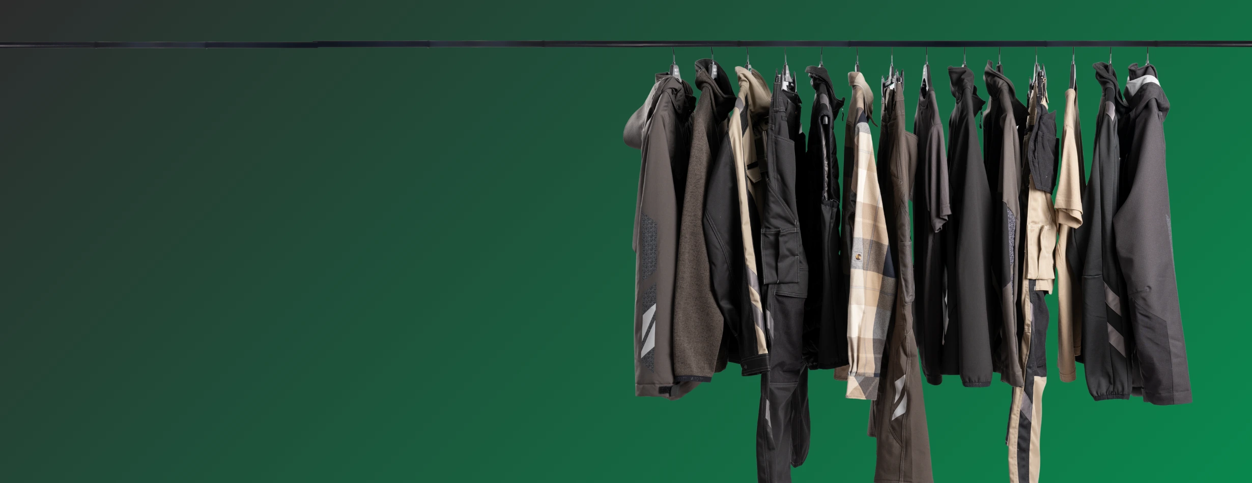 Mehrere Arbeitsjacken in dunklen und neutralen Farbtönen hängen ordentlich auf einer Kleiderstange vor einem einfarbig grünen Hintergrund.
